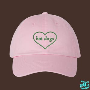 May include: A light pink baseball cap featuring an embroidered green heart outline on the front panel. Inside the heart, the text "hot dogs" is stitched in green lowercase letters. The cap has a structured crown, a curved brim, and a button top.