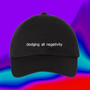 Pode incluir: Um boné de beisebol preto com o texto bordado em branco "dodging all negativity" centralizado no painel frontal. O boné possui uma coroa estruturada, uma aba curva e um botão no topo, proporcionando um visual clássico e definido.
