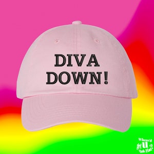 Pode incluir: Boné de beisebol rosa claro com o texto "DIVA DOWN!" bordado em preto. O boné tem uma aba curva e um botão na parte superior. O fundo é um gradiente de rosa, amarelo, verde e laranja.