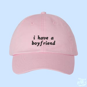 May include: Pink baseball cap with the text "i have a boyfriend" embroidered in black.