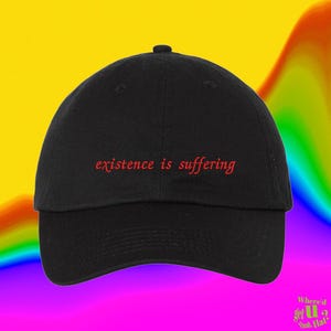 May include: A black baseball cap featuring the text "existence is suffering" embroidered in red thread across the front panel. The cap has a curved brim, a button on the top centre, and stitched eyelets for ventilation on the crown panels.