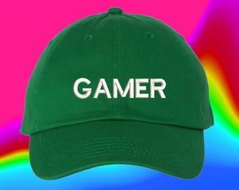 May include: A forest green baseball cap featuring the word GAMER embroidered in white capital letters across the front center. The cap has a structured crown, a curved brim, and visible stitching details along the panels and the brim edge.