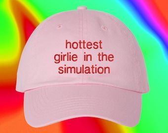 May include: A light pink baseball cap featuring the embroidered red text "hottest girlie in the simulation" centered on the front panel. The cap has a structured crown, a curved brim, and a button at the top, with visible stitching details.