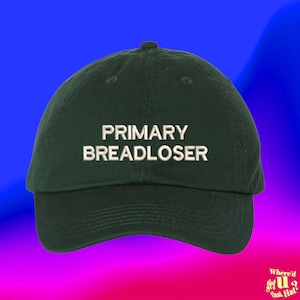 May include: A dark green baseball cap featuring the embroidered white text PRIMARY BREADLOSER centred on the front panel. The cap has a structured crown, a curved brim, and stitched eyelets for ventilation, presenting a clean and simple design.