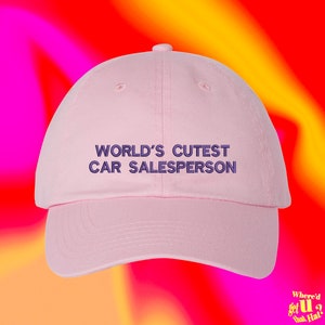 Puede incluir: Gorra de béisbol rosa con el texto "WORLD'S CUTEST CAR SALESPERSON" bordado en morado.