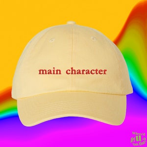 Puede incluir: Una gorra de béisbol amarilla con el texto "main character" bordado en rojo.