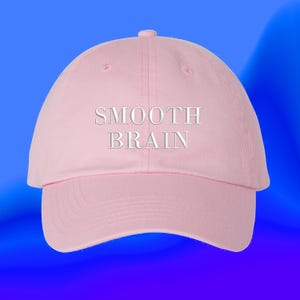 May include: A light pink baseball cap featuring the text SMOOTH BRAIN embroidered in white capital letters across the front panel. The cap has a structured crown, a curved brim, and a button at the top center of the dome.