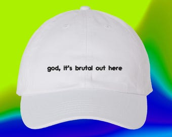 May include: A white baseball cap featuring the black embroidered text "god, it's brutal out here" centered on the front panel. The cap has a structured crown, a curved brim, and a button at the top, presenting a clean and simple design.