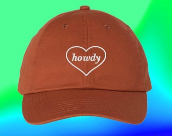 May include: A rust-coloured baseball cap featuring an embroidered white heart outline on the front panel. Inside the heart, the word howdy is stitched in a white cursive font. The cap has a curved brim and a button on the top centre.