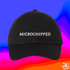 May include: A black baseball cap featuring the word MICROCHIPPED embroidered in white capital letters across the front center. The hat has a structured crown, a curved brim, and a button at the top center, showcasing a clean and simple design.