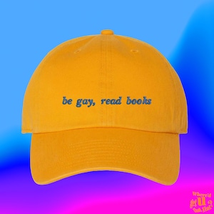 Peut inclure: Une casquette de baseball jaune avec le texte "be gay, read books" brodé en bleu.