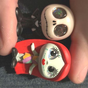 Puede incluir: Tres figuras de personajes de Pesadilla antes de Navidad. Jack Skellington, con un traje negro, y Sally, con un vestido de retazos, están representados. Las figuras tienen ojos brillantes y están sobre un fondo oscuro.