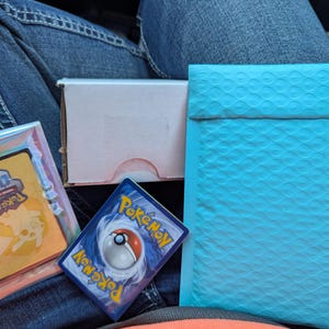 Op de afbeelding: Een verzameling Pokémon-ruilkaarten, waaronder een holografische kaart in een beschermhoes, een kaart met het Pokémon-logo en een lichtblauwe bubbelenvelop. Er is ook een kleine, open kartonnen doos aanwezig.