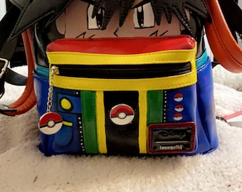 May include: A black, red, yellow, green, and blue backpack with a cartoon face of a character wearing a red and white baseball cap. The backpack has a zippered pocket and a small charm attached to it. The charm is a red and white Pokeball.