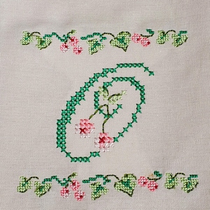 Può includere: Un ricamo a punto croce su tessuto beige chiaro con una lettera O centrale formata da viticci verdi e piccoli fiori rosa. La lettera è incorniciata da bordi orizzontali di foglie verdi e bacche rosa nella parte superiore e inferiore.