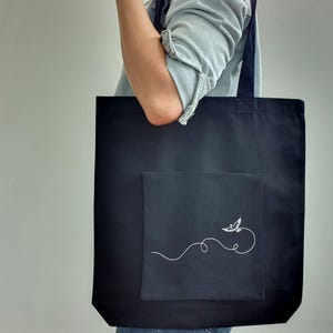May include: Black tote bag with a square pocket featuring a white embroidered bird design. The bag has a simple, minimalist aesthetic, with a long strap for carrying over the shoulder. The bag is made of a sturdy fabric.