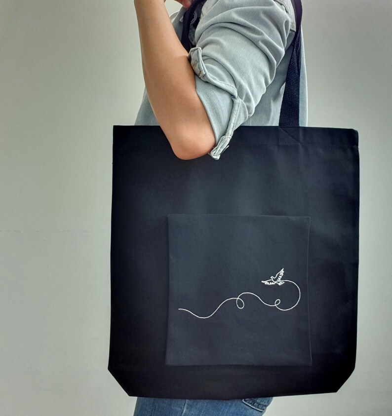 May include: Black tote bag with a square pocket featuring a white embroidered bird design. The bag has a simple, minimalist aesthetic, with a long strap for carrying over the shoulder. The bag is made of a sturdy fabric.