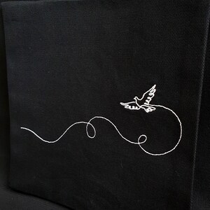 May include: Black fabric with a white embroidered design of a dove in flight. The dove is depicted with outstretched wings, trailing a curved, looping line. The embroidery is detailed and delicate, creating a striking contrast against the dark background.