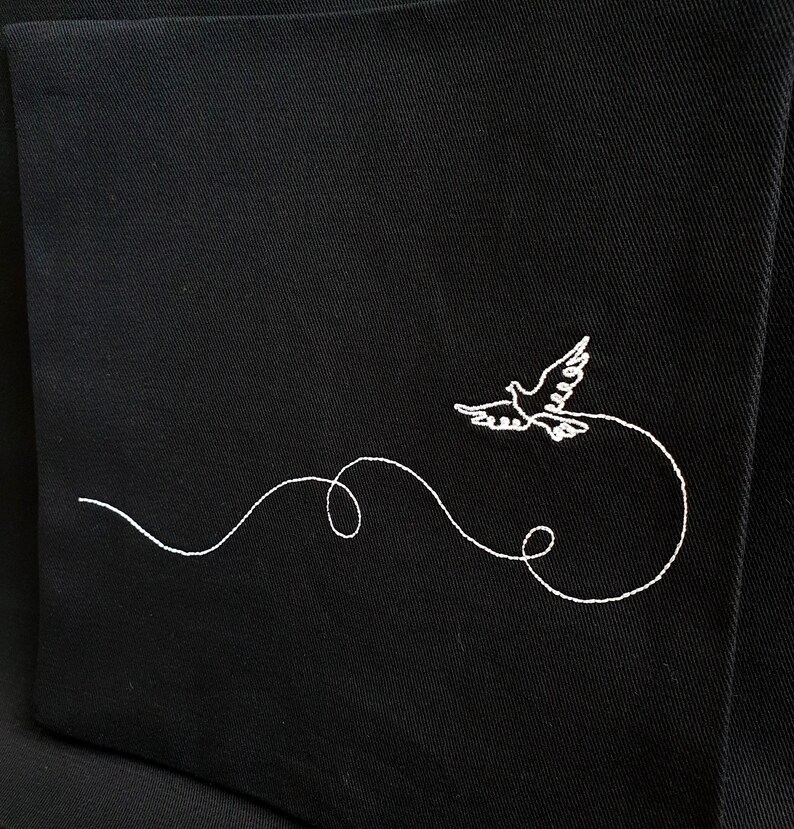 May include: Black fabric with a white embroidered design of a dove in flight. The dove is depicted with outstretched wings, trailing a curved, looping line. The embroidery is detailed and delicate, creating a striking contrast against the dark background.