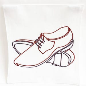 May include: White pillow with an embroidered design of a dress shoe in a minimalist style. The shoe outline is in dark red and navy blue thread, with the laces and sole details visible.