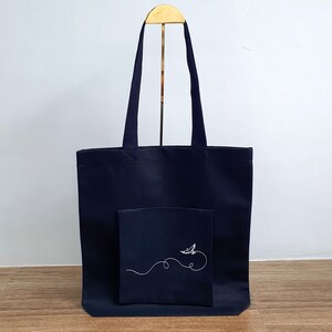 May include: A navy blue tote bag with a matching pocket featuring a white embroidered bird design. The bag has two navy blue handles and is displayed on a gold stand. The tote bag is made of a durable fabric.