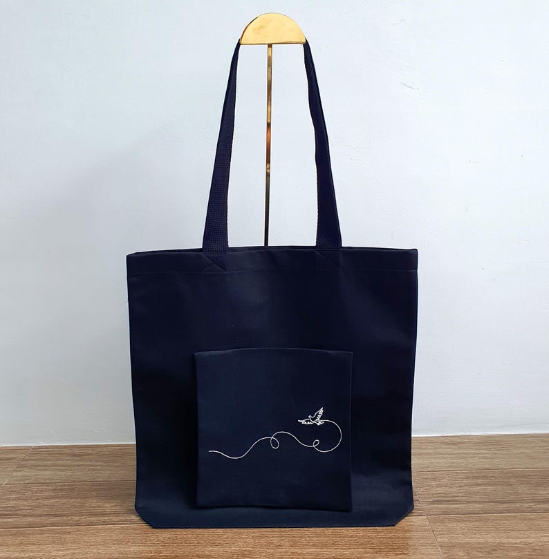 May include: A navy blue tote bag with a matching pocket featuring a white embroidered bird design. The bag has two navy blue handles and is displayed on a gold stand. The tote bag is made of a durable fabric.
