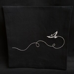 May include: Black fabric item with a white embroidered design of a dove in flight. The dove is depicted with outstretched wings, trailing a curved line. The design is centered on the fabric, creating a simple yet elegant aesthetic.