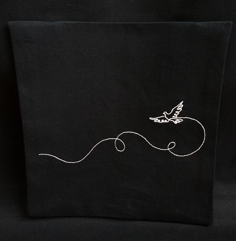 May include: Black fabric item with a white embroidered design of a dove in flight. The dove is depicted with outstretched wings, trailing a curved line. The design is centered on the fabric, creating a simple yet elegant aesthetic.