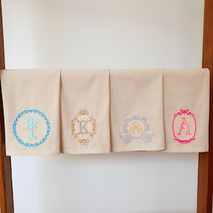 Puede incluir: Cuatro toallas de lino beige con diseños bordados. Cada toalla presenta una inicial diferente: T, K, R y A. Los colores del bordado incluyen azul, dorado, gris y rosa. Las toallas están colgadas en un estante de madera.