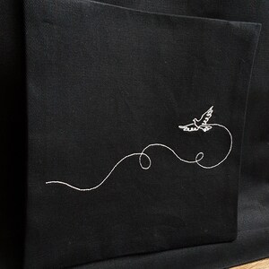May include: Black tote bag with a white embroidered design of a flying dove and a curved line. The dove is depicted in flight, with its wings spread. The design is on the front of the bag, which has a simple, minimalist style.