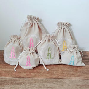 Puede incluir: Bolsas beige con cordón y bordado de la letra "A" en varios colores, como rosa, verde, amarillo y azul. Cada bolsa tiene un borde blanco con diseño de encaje alrededor de la letra. Las bolsas están dispuestas sobre una superficie de madera.