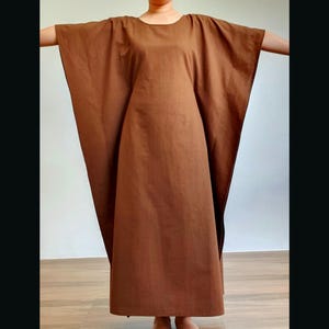 May include: A solid brown kaftan dress featuring a round neckline and wide, flowing sleeves that extend from the shoulders. The garment has a loose, floor-length silhouette with a simple, unadorned design and a smooth, matte fabric texture.