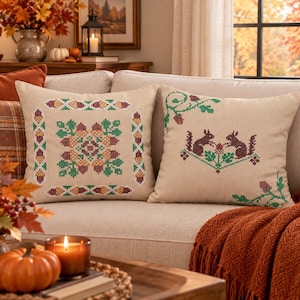 May include: Two square beige throw pillows featuring cross-stitch embroidery. The left pillow displays a central geometric pattern of acorns and leaves. The right pillow shows two brown squirrels facing each other with green foliage and acorns.