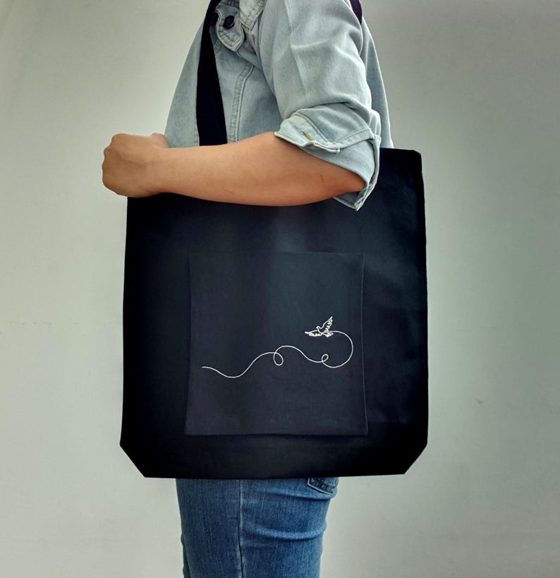 May include: Black tote bag with a white embroidered design of a bird in flight. The tote bag has a large front pocket and long straps. The bag is being held by a person wearing a denim jacket and blue jeans.
