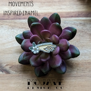 May include: Gold enamel pin with a hand holding a white flower and the text "Feel Something". The pin is on a purple succulent plant.