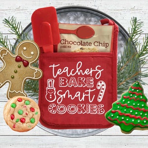 May include: Red oven mitt with the text "teachers bake smart cookies" in white. A gingerbread man cookie, a chocolate chip cookie mix bag, a red spatula, and a green Christmas tree cookie are arranged around the oven mitt.