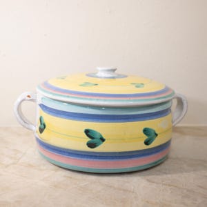 May include: A ceramic casserole dish with a lid. The dish features yellow, blue, and pink horizontal stripes. Teal heart-shaped designs are scattered around the dish. The handles and lid knob are white.