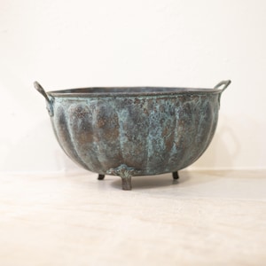 Puede incluir: Un cuenco de metal envejecido, de estilo antiguo, con un diseño acanalado y un acabado patinado. El cuenco tiene dos pequeñas asas y descansa sobre tres patas decorativas. El color es una mezcla de bronce y verde azulado. Un objeto decorativo.