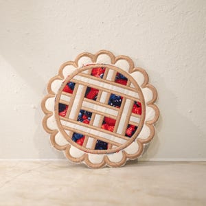 Könnte beinhalten: Ein runder Stoffuntersetzer, der einem Beerenkuchen ähnelt. Der Untersetzer hat ein Gitterkrustendesign mit einem beigen Rand und einem gewellten Rand. Die Füllung ist eine Mischung aus roten Erdbeeren und blauen Blaubeeren.