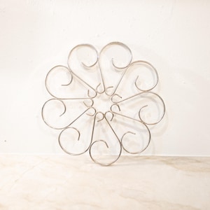 May include: A silver metal trivet with a floral design. The trivet has a circular shape with curved, heart-like elements radiating from a central point. The metal has a shiny finish.