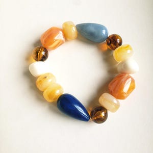 Peut inclure: Un bracelet élastique composé de perles assorties de formes et de tailles variées. Les perles présentent un mélange de finitions unies, translucides et marbrées dans des tons orange, jaune, blanc, bleu foncé, bleu clair et marron foncé.