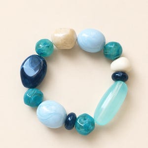 Peut inclure: Un bracelet élastique composé de perles lisses et polies de formes et de tailles variées. Les perles présentent une palette de couleurs bleu clair, bleu marine foncé, sarcelle, turquoise et crème, avec des finitions marbrées ou translucides.