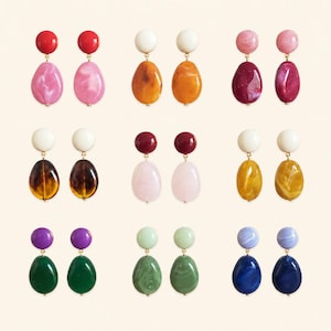 May include: Nine pairs of drop earrings arranged in a grid, each featuring a round stud top and a larger, irregular oval-shaped drop pendant. The earrings display a variety of solid and marbled colors, including red, pink, orange, white, burgundy, yellow, purple, green, and blue tones.