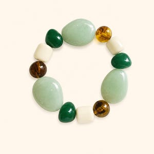 May include: A stretch bracelet featuring a variety of smooth, polished beads in shades of light mint green, deep forest green, cream, and translucent amber brown. The beads are arranged in a circular pattern, alternating between large oval shapes and smaller round or cylindrical forms.