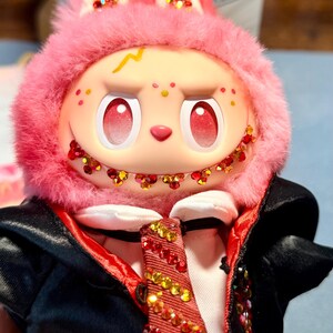 May include: A plush doll with a pink fuzzy hood featuring red and gold glitter accents on the ears and teeth. The doll has a pale face with red gradient eyes, a yellow lightning bolt mark, and wears a black jacket with a red lining and a red tie.