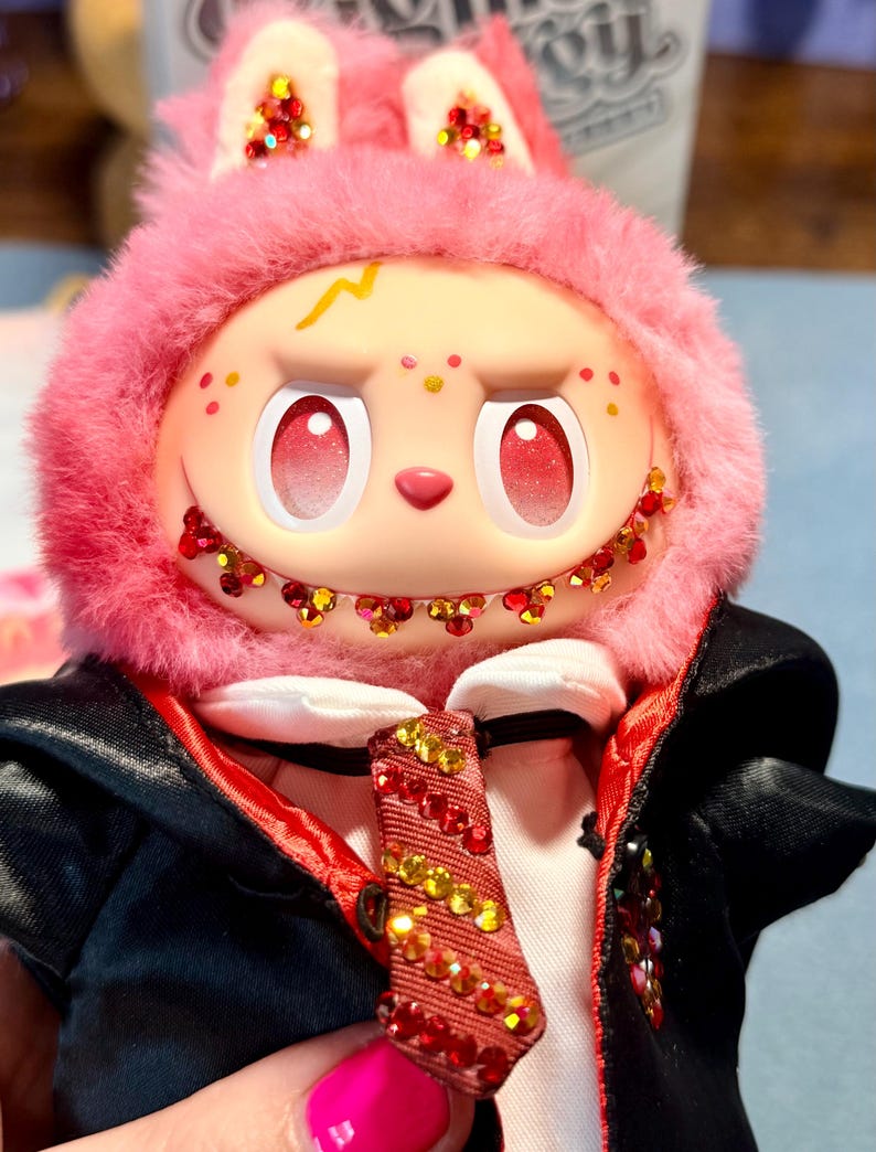 May include: A plush doll with a pink fuzzy hood featuring red and gold glitter accents on the ears and teeth. The doll has a pale face with red gradient eyes, a yellow lightning bolt mark, and wears a black jacket with a red lining and a red tie.