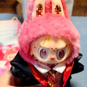 May include: A collectible vinyl figure wearing a pink fuzzy rabbit hood with embedded crystals on the ears. The figure has a pale face, a yellow lightning bolt mark on the forehead, and large red eyes. It wears a black jacket with a red lining and a crystal-embellished tie.