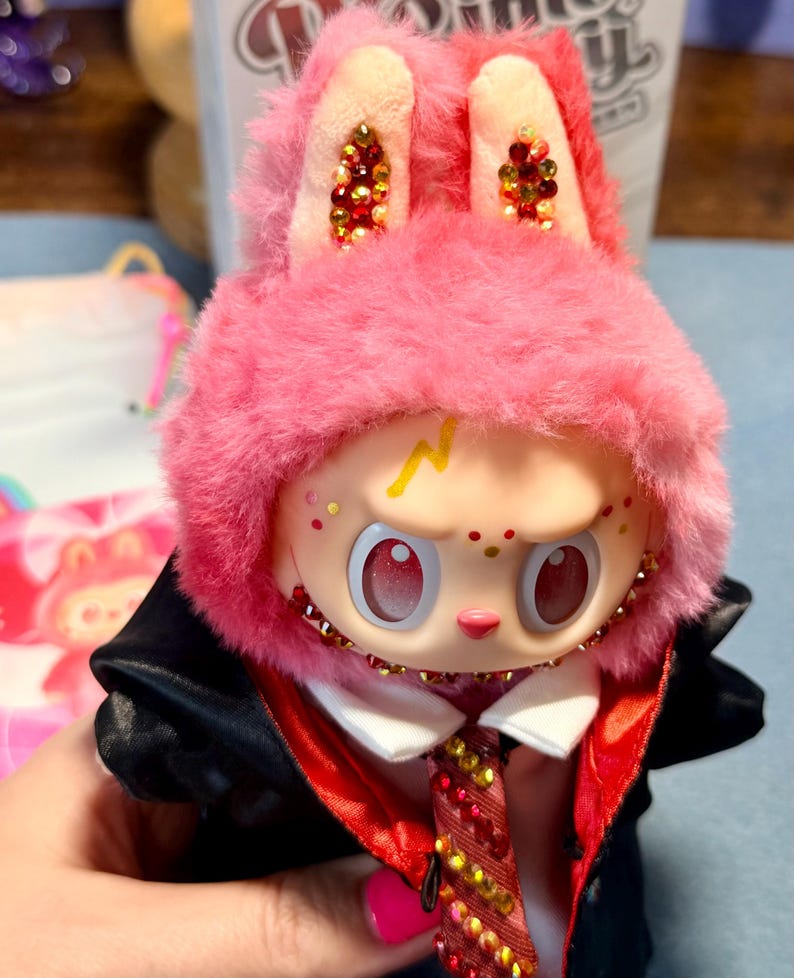 May include: A collectible vinyl figure wearing a pink fuzzy rabbit hood with embedded crystals on the ears. The figure has a pale face, a yellow lightning bolt mark on the forehead, and large red eyes. It wears a black jacket with a red lining and a crystal-embellished tie.