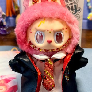 May include: A plush toy figure with pink fur, large eyes, and a yellow lightning bolt mark on its forehead. It wears a white shirt, a red and yellow striped tie adorned with crystals, and a black robe with a red lining. The face features crystal accents.