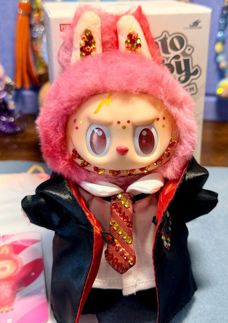 May include: A plush toy figure with pink fur, large eyes, and a yellow lightning bolt mark on its forehead. It wears a white shirt, a red and yellow striped tie adorned with crystals, and a black robe with a red lining. The face features crystal accents.